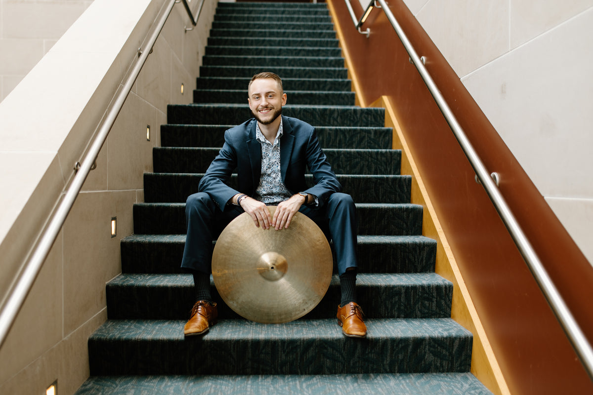 Ryan Barski seated on a staircase in a navy suit, smiling, with his hands resting on a ride cymbal